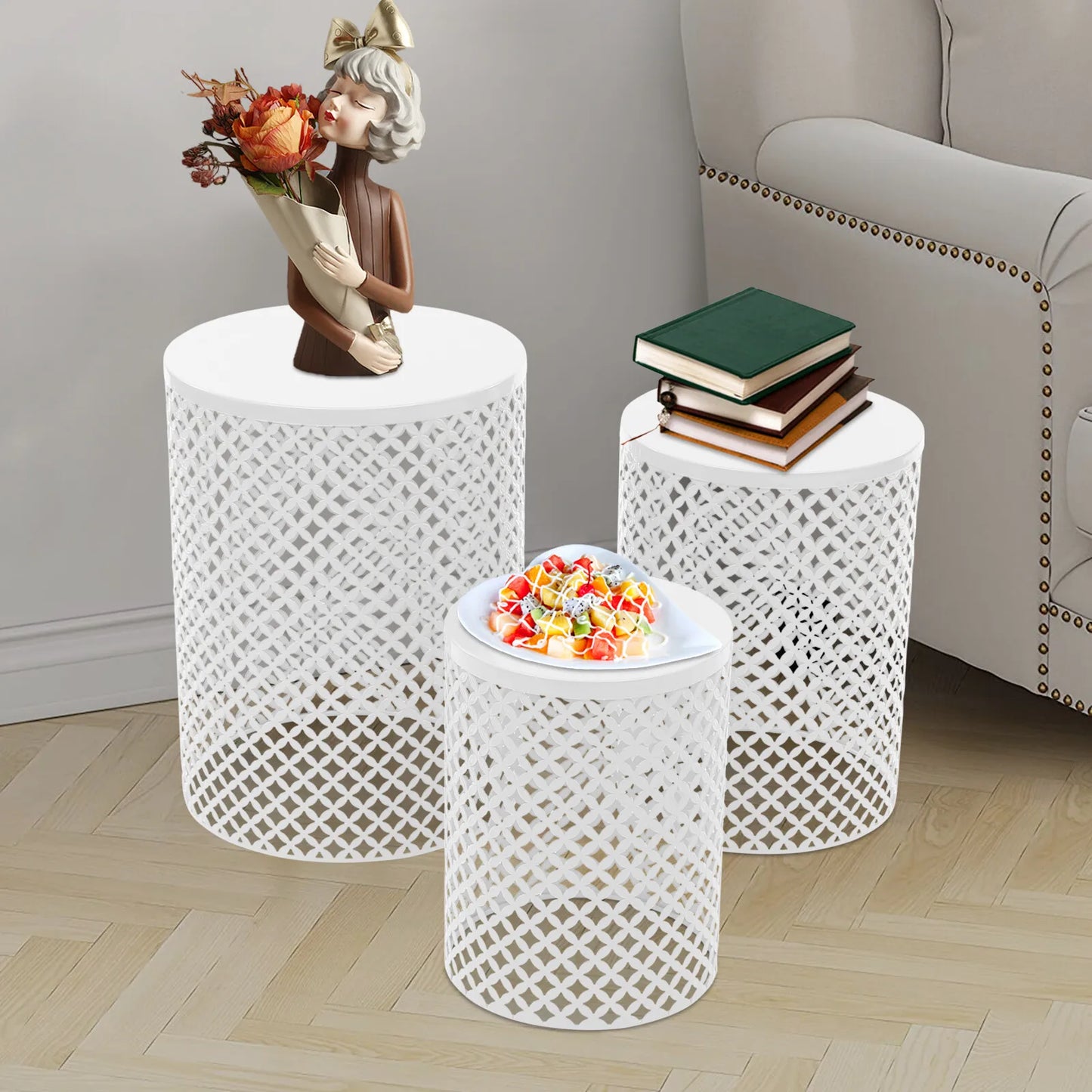 Three white metal lattice end tables; tallest holds bouquet statue, middle stacks books, smallest holds candy dish