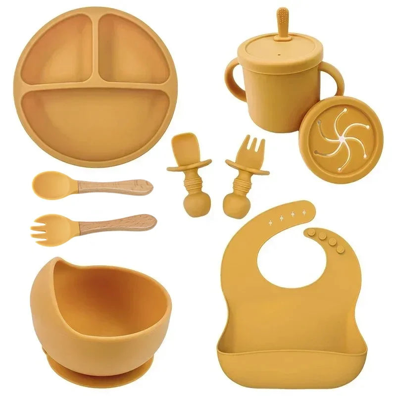 Food-grade silicone baby feeding set by S And B Enterprise; mustard yellow suction plate, straw cup, bib, bowl, and wooden-handled spoons