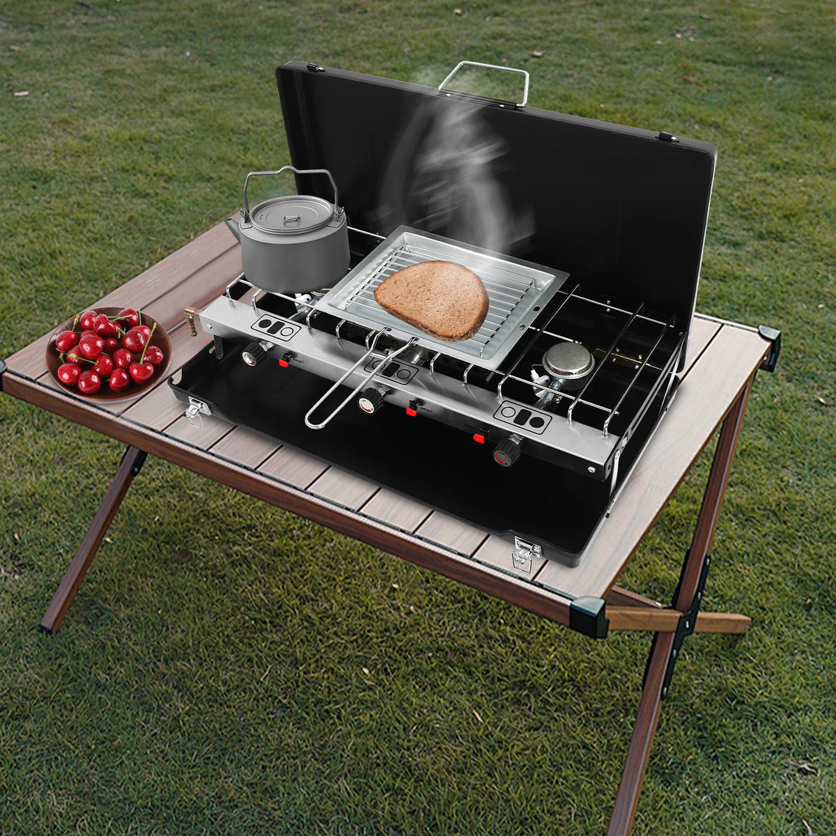 Portable table-top 3-burner camping stove, black with stainless rack; kettle, bread slice, and cherries nearby
