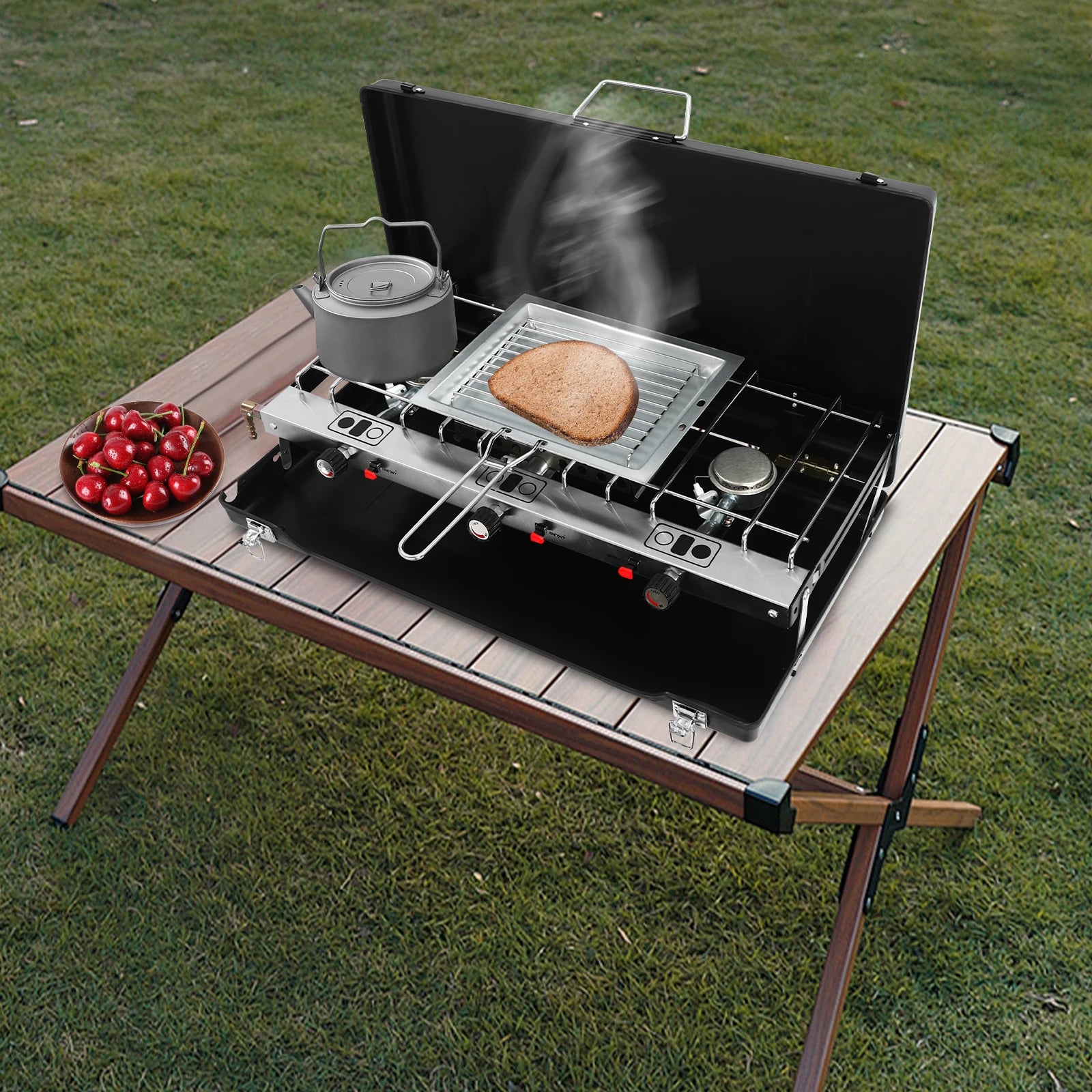 Portable table-top 3-burner camping stove, black with stainless rack; kettle, bread slice, and cherries nearby
