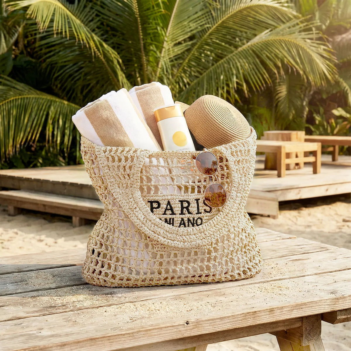 Natural straw woven beach tote with Paris Milano branding; towels, sunscreen bottle, hat and sunglasses visible