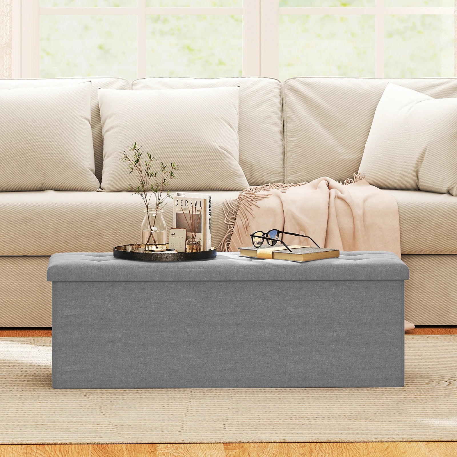 Fabric folding storage ottoman in dark grey placed in front of a beige sofa, with a tray featuring a small plant and decorative items.