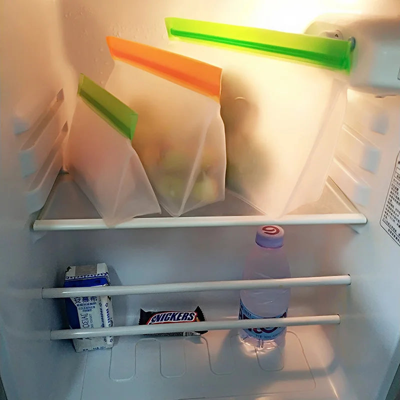 Three reusable silicone PEVA stand-up freezer bags with leakproof tops in orange and green, on a fridge shelf.