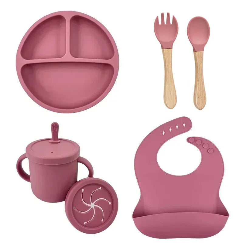 Pink silicone baby feeding set with divided plate, wooden-handled spoon and fork, sippy cup with lid, and silicone bib